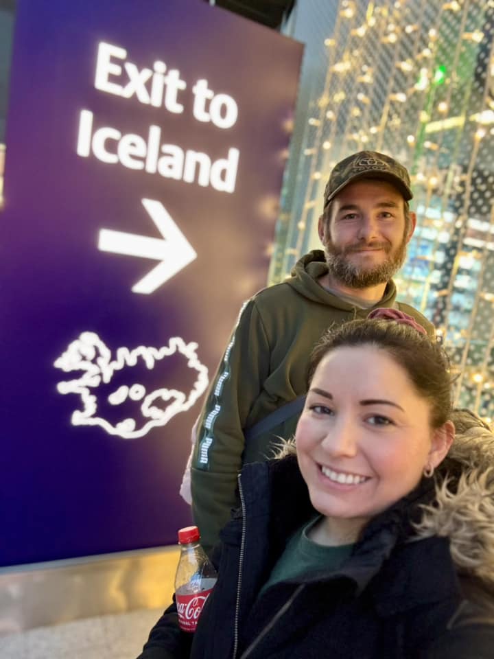 A selfie of a smiling woman in a puffer jacket and a man in a camo hat at Keflavík Airport, standing next to a large purple sign with an arrow and text that reads "Exit to Iceland."