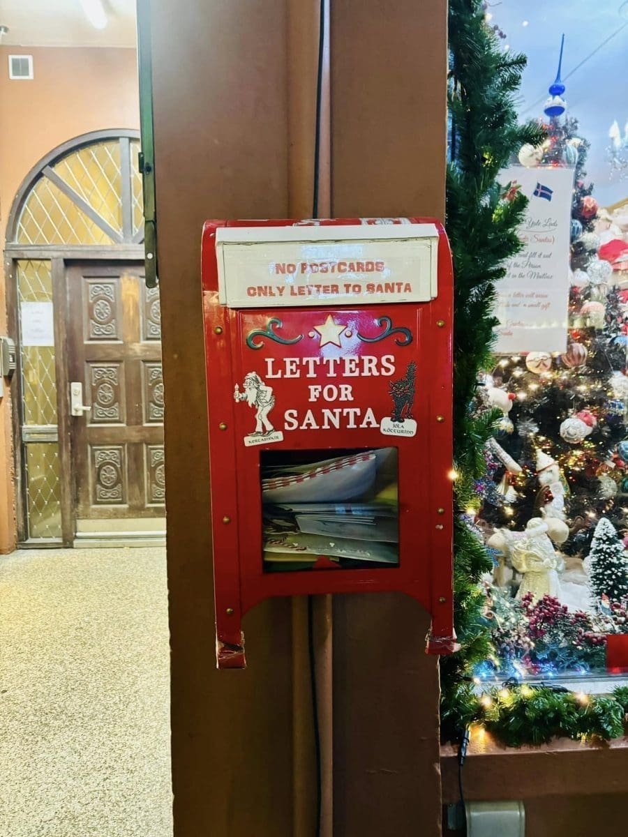 A bright red vintage-style mailbox with white lettering that reads "Letters for Santa" and a sign stating "No Postcards, Only Letter to Santa," placed next to a decorated Christmas tree.
