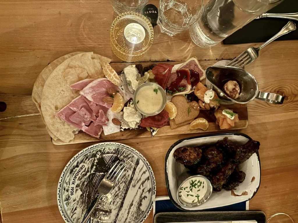 A top-down view of a rustic wooden table featuring a large charcuterie board with smoked salmon, prosciutto, and cheeses, a bowl of dark glazed wings, and a decorative plate with "Grindavík 1926" written on it.
