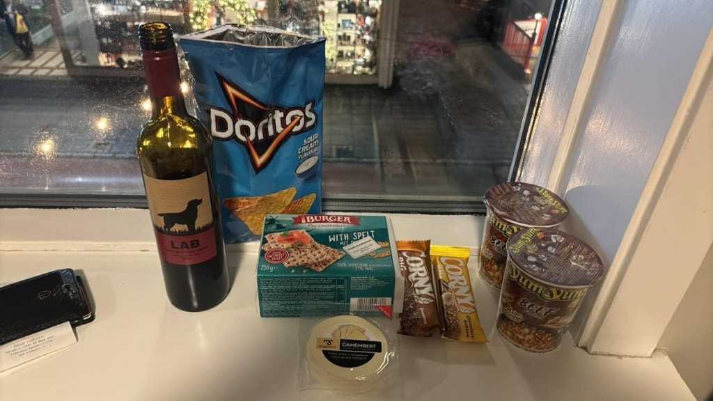 A hotel windowsill stocked with travel snacks including a bottle of red wine, Doritos, cheese, crackers, and instant noodles, with a view of a festive Reykjavik street outside.