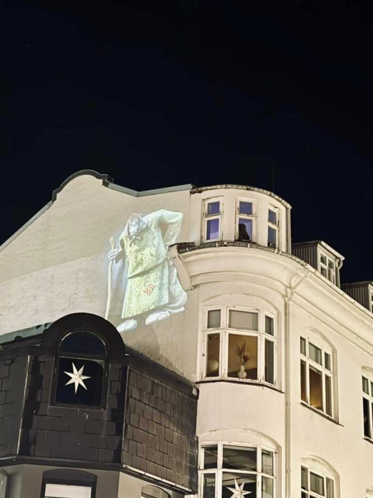 A large, glowing projection of a traditional Icelandic "Yule Lad" goblin holding a staff, illuminated on the side of a white historic building at night.