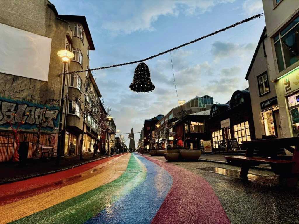 The famous Rainbow Road in downtown Reykjavik, Iceland, decorated with a giant hanging Christmas bell and glowing holiday lights under a soft evening sky.