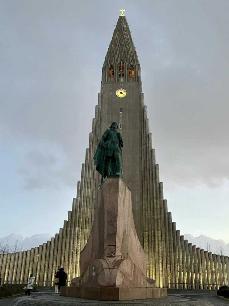 A low-angle shot of the iconic Hallgrímskirkja church in Reykjavik, featuring its unique stepped concrete facade and the statue of Leif Erikson standing prominently in front.