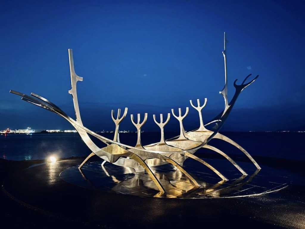 The Sun Voyager (Sólfar) stainless steel sculpture glowing under blue night lights on the Reykjavik waterfront, with the dark ocean and city lights in the background.