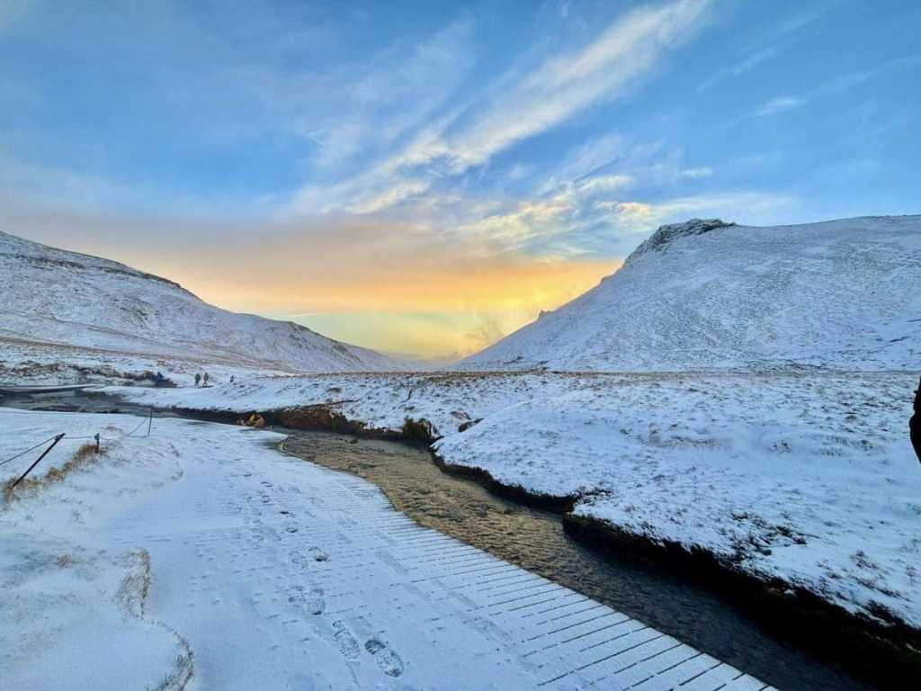 A view from a snowy mountain trail in Reykjadalur, Iceland, showing steam rising from geothermal vents in the valley below against a backdrop of vast, snow-covered mountain ranges under a clear sky.
