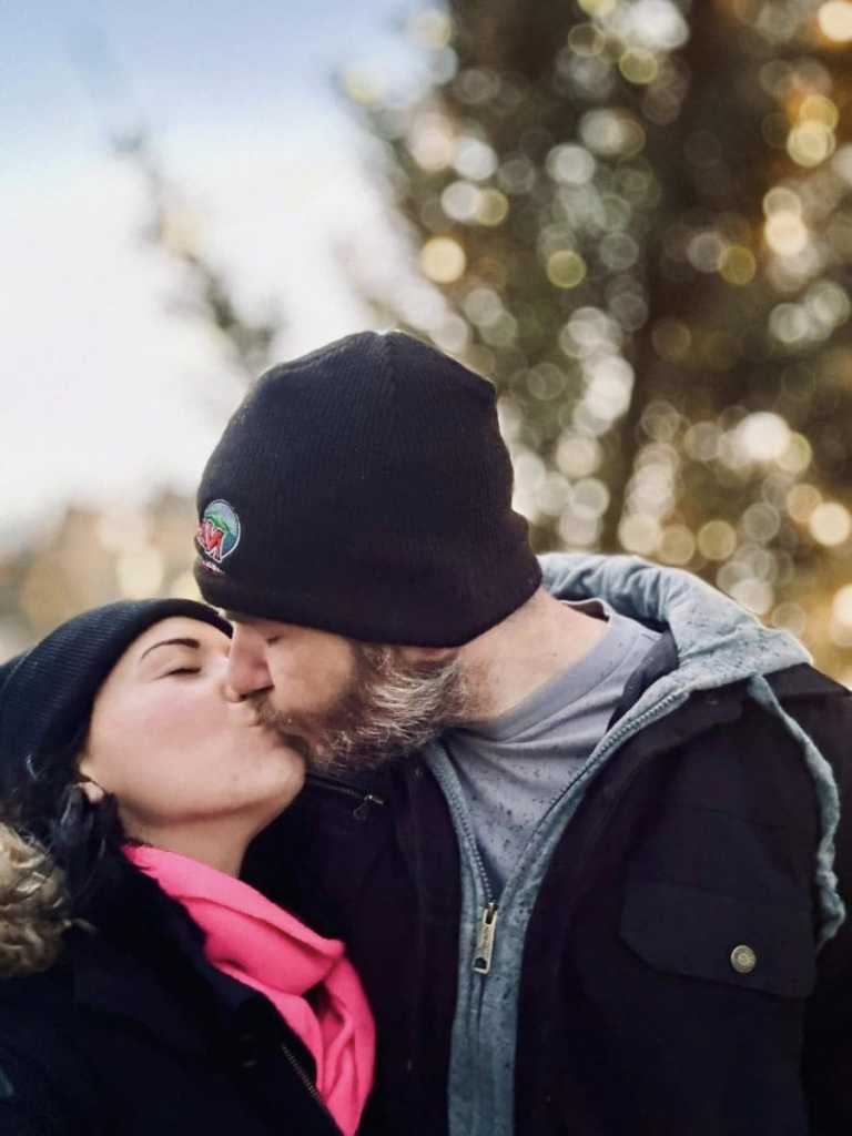 A close-up, romantic photo of a couple wearing beanies and winter coats sharing a kiss in front of a background of blurred holiday lights.