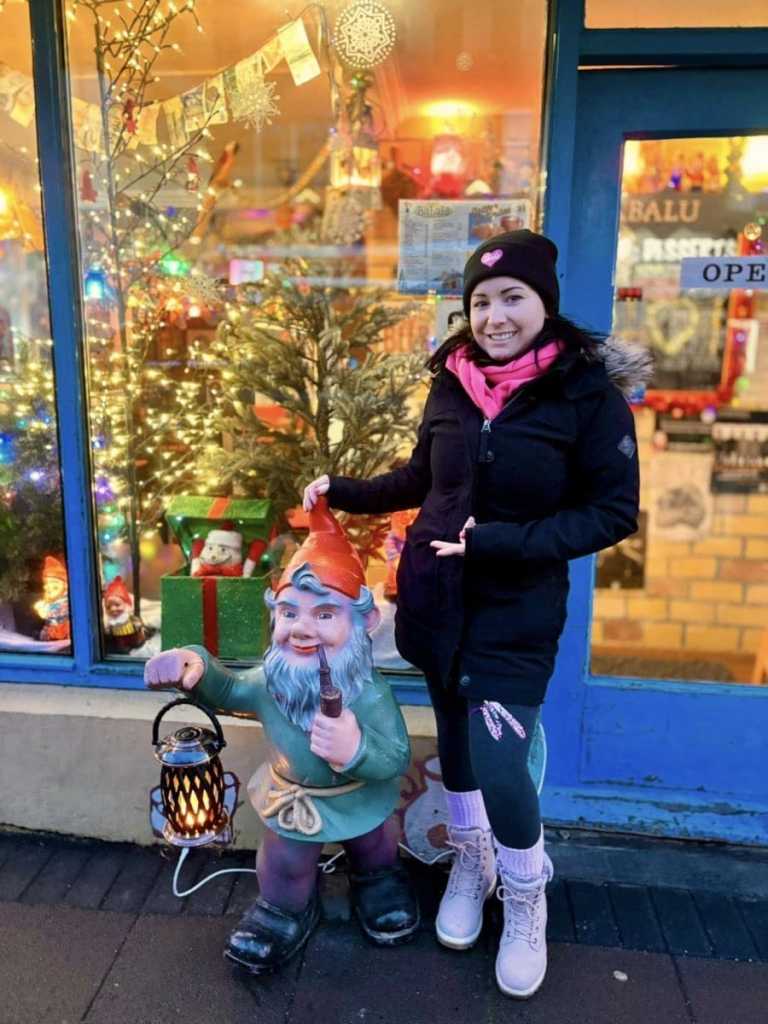 A woman in a pink scarf and black parka posing next to a colorful statue of a bearded Yule Lad carrying a lantern outside a decorated shop window.