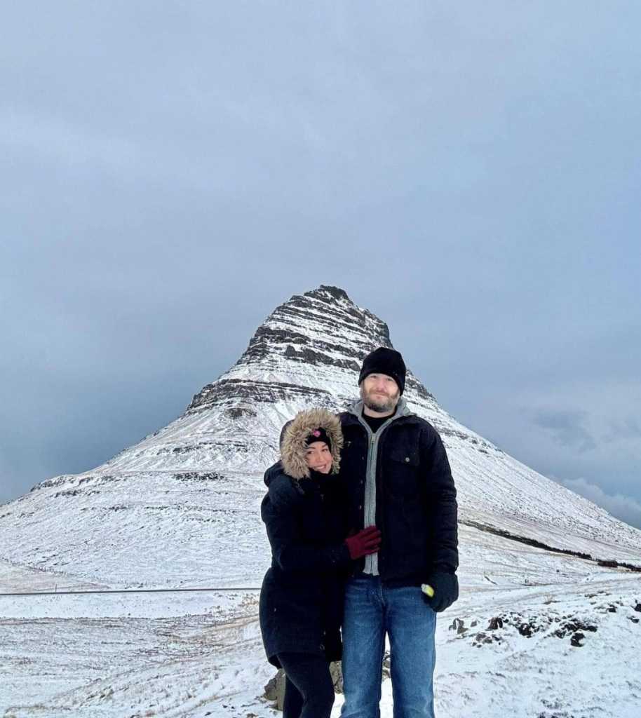A happy couple standing closely together in front of the iconic Kirkjufell mountain in Iceland. The mountain towers behind them with its distinct cone shape covered in patches of snow under a soft, overcast sky. Both are dressed in warm winter gear—a woman in a black parka with a fur-lined hood and maroon gloves, and a man in a black jacket, beanie, and blue jeans—smiling as they enjoy the majestic scenery.