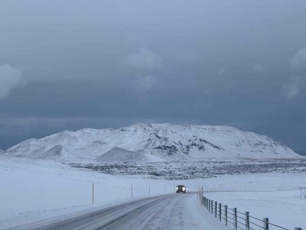 A view from a car driving on a narrow, snow-covered road through a vast, white Icelandic tundra. In the distance, a massive, jagged mountain covered in heavy snow looms under a dark, moody grey sky, with another vehicle's headlights visible as it approaches from the opposite direction.