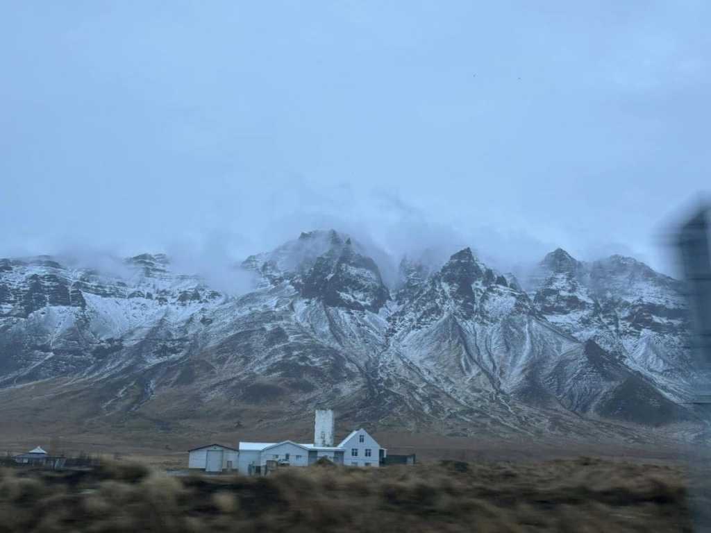 A view from the passenger seat of a car driving along a paved highway in Iceland towards Akranes. The road stretches toward snow-capped mountains under a vast, bright sky, with a rugged, grassy landscape lining the side of the road.