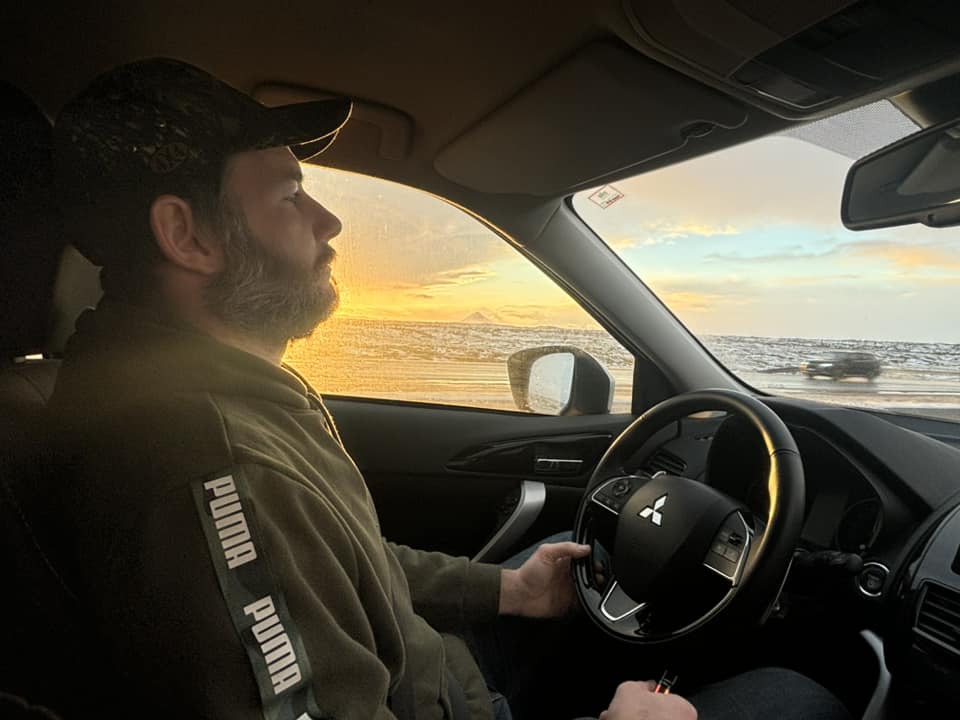 A man in a Puma hoodie and a camo hat driving a Mitsubishi rental car through the Icelandic landscape. The golden morning sun is shining brightly through the window, illuminating the snow-covered fields outside.