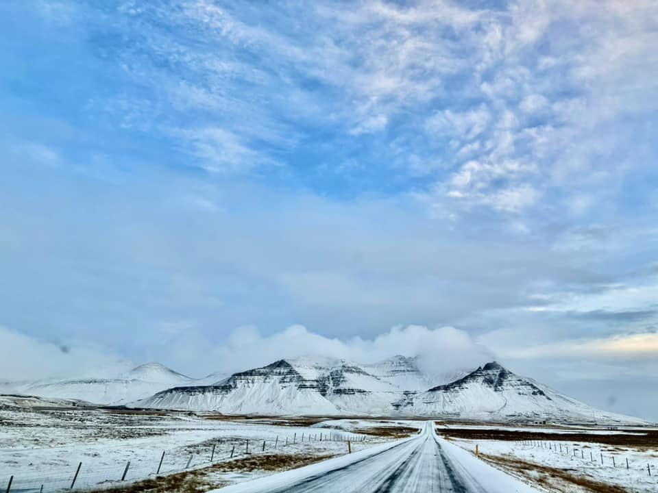 A point-of-view shot looking down a long, straight, two-lane road in Iceland, covered in light snow and ice. The road leads directly toward a massive range of jagged, white-capped mountains that disappear into low-hanging clouds. The sky above is a bright, clear blue with soft white clouds, and the surrounding fields are a mix of frost-covered grass and dark volcanic earth.