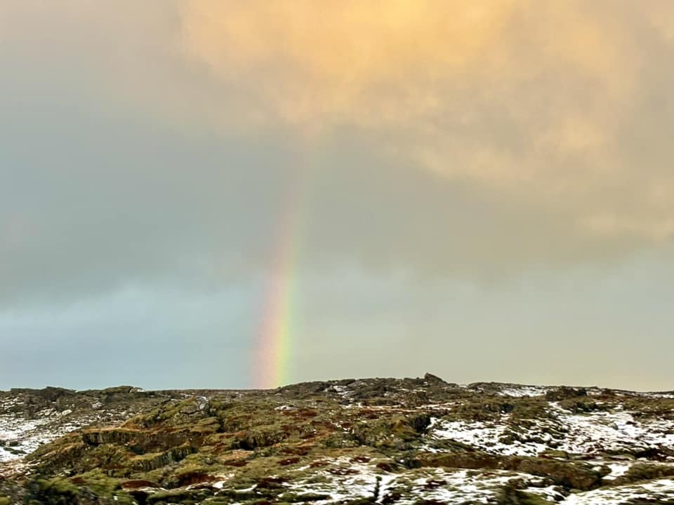 A view from a vehicle driving toward Keflavík Airport through rugged, moss-covered lava fields partially dusted with snow. In the background, a dark, heavy storm cloud recedes, revealing a bright, vibrant rainbow arching against a soft, sunlit orange and yellow sky. The foreground is filled with the dark volcanic rock formations typical of the Reykjanes Peninsula.