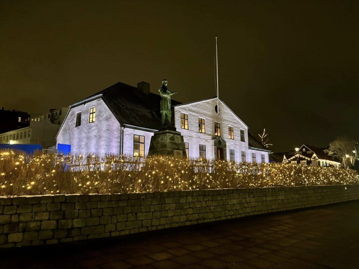 A historic white two-story building in Reykjavik illuminated at night, featuring a statue on a pedestal in front and a hedge glowing with warm yellow Christmas lights.