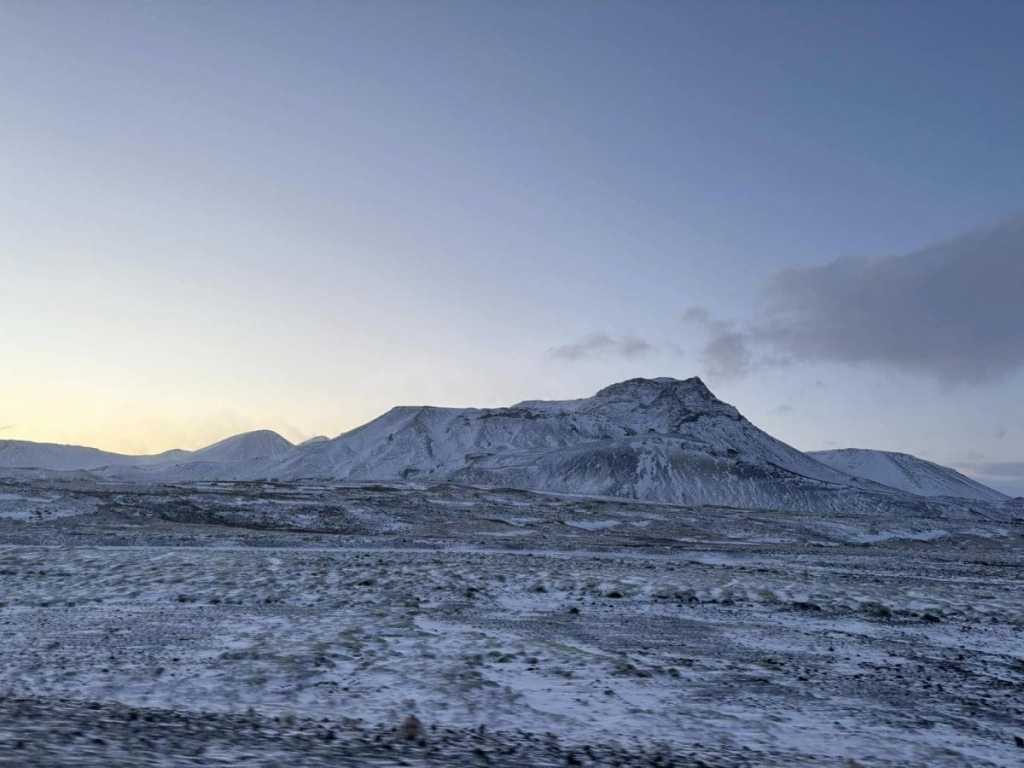 A wide panoramic view of snow-covered Icelandic mountains under a pale blue sky at dawn, with the white and grey tundra stretching across the foreground.