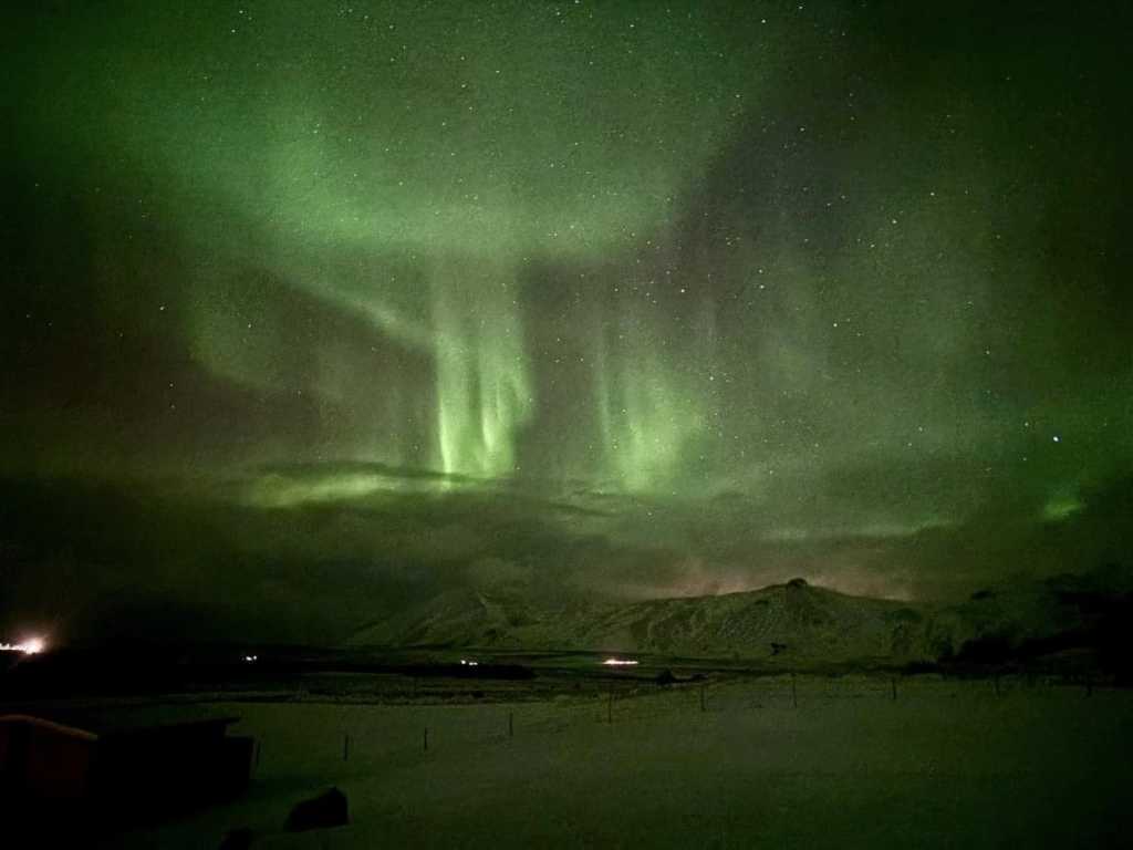 A breathtaking night view from a large window in Akranes, Iceland, looking out over a snow-covered landscape. Faint, ethereal green Northern Lights (Aurora Borealis) dance across the dark sky above a massive mountain range with jagged, snowy peaks, with a calm body of water reflecting the soft light at the base.