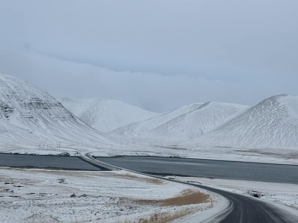 A wide-angle landscape shot taken from a road winding through a snow-covered mountain valley in Iceland. The dark asphalt road curves around a calm, dark blue body of water, leading toward distant, smooth white mountains under a thick, overcast sky. The foreground shows patches of brown winter grass peeking through the snow.