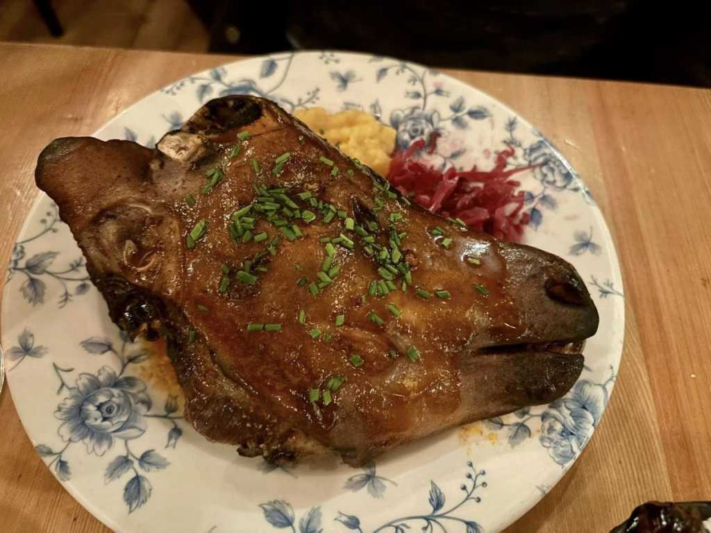 A close-up of a traditional roasted Icelandic sheep's head served on a blue and white floral plate, garnished with fresh chives and served with a side of mashed turnips and red cabbage.