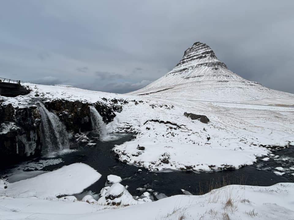 A wide, scenic view of Kirkjufellsfoss waterfall in Iceland during winter. Two streams of water cascade into a partially frozen pool in the foreground, surrounded by snow-covered rocks. In the background, the distinctive, cone-shaped Kirkjufell mountain rises majestically against a moody, overcast sky, creating a powerful and serene winter landscape.