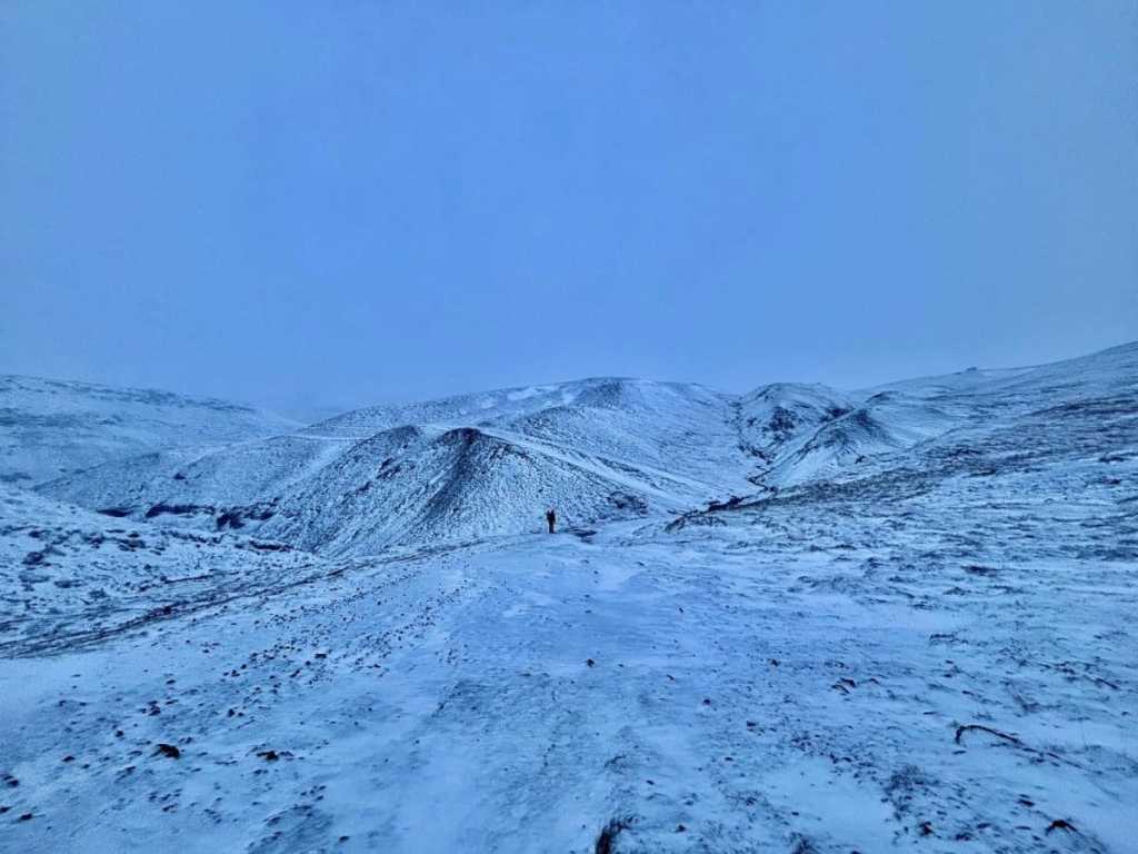 A person in a black patterned winter jacket and a grey beanie stands on a winding, snow-covered mountain trail overlooking a vast geothermal valley in Iceland, with steam rising from the ground in the distance under a pale blue sky.