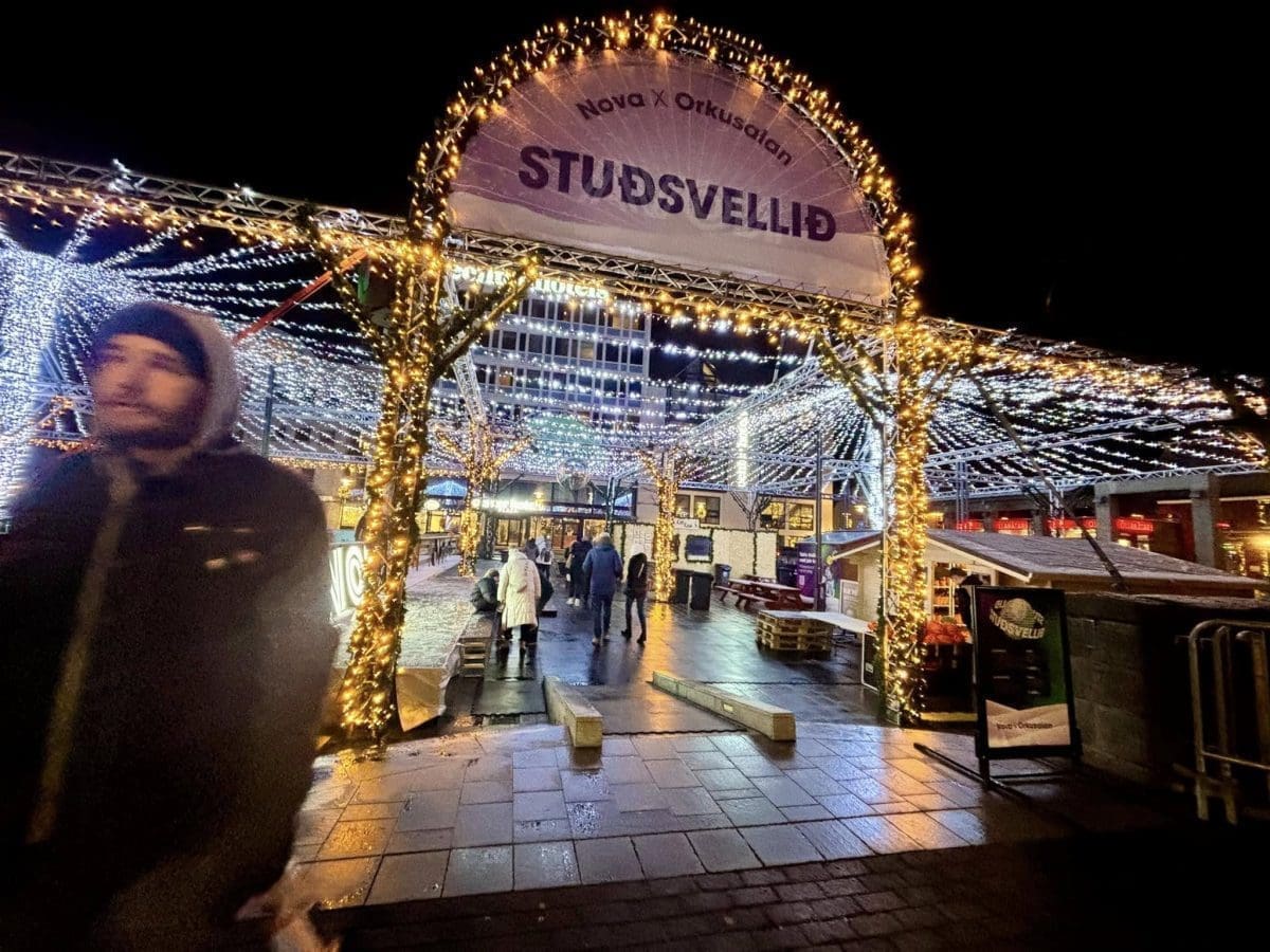 An outdoor Christmas market at night with a lit-up entrance sign that reads "STUÐSVELLIÐ," featuring rows of twinkling lights and people walking through a festive plaza.