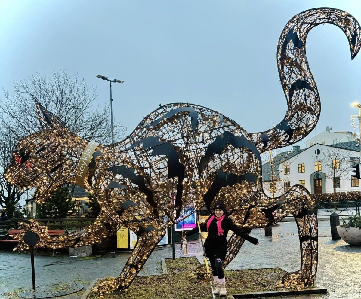 A woman standing next to a massive, towering holiday sculpture of a cat made of woven branches and hundreds of sparkling Christmas lights in downtown Reykjavik.