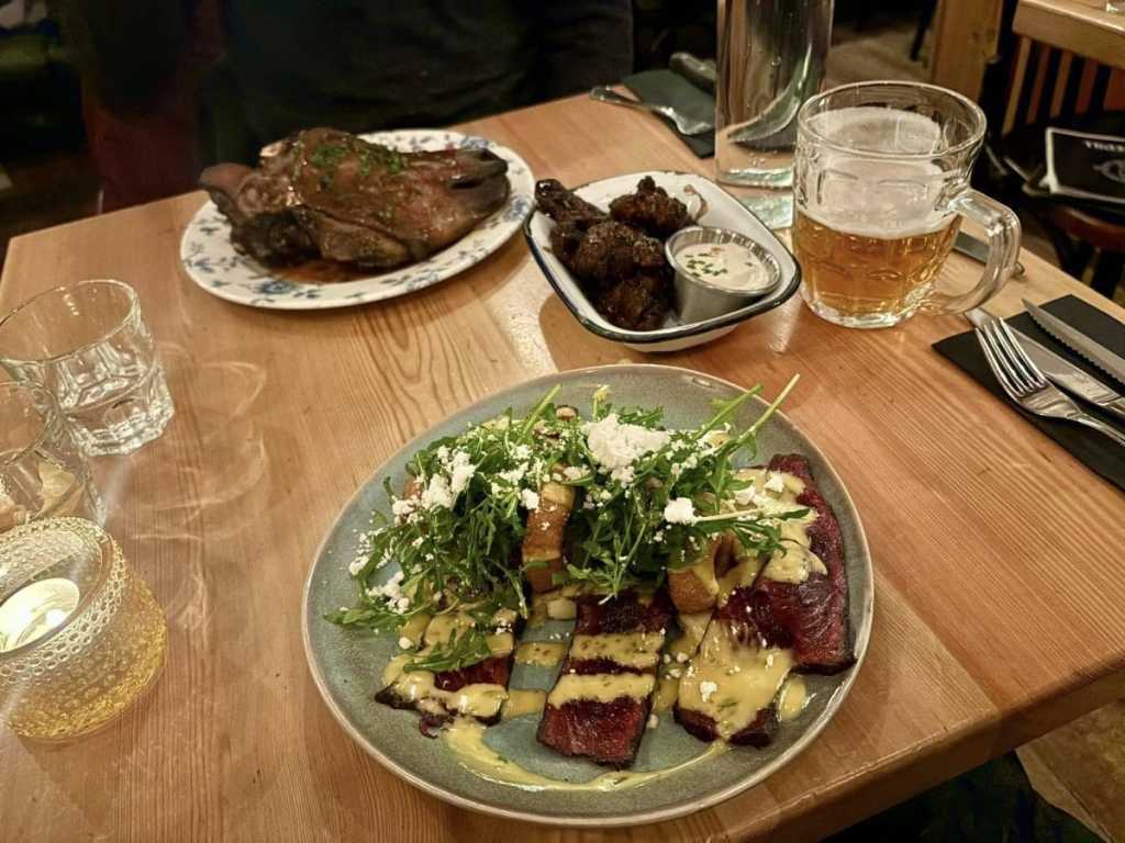 A restaurant table featuring a plate of thinly sliced whale meat topped with arugula and white cheese crumbles in the foreground, with a traditional roasted sheep's head and a glass of beer in the background.
