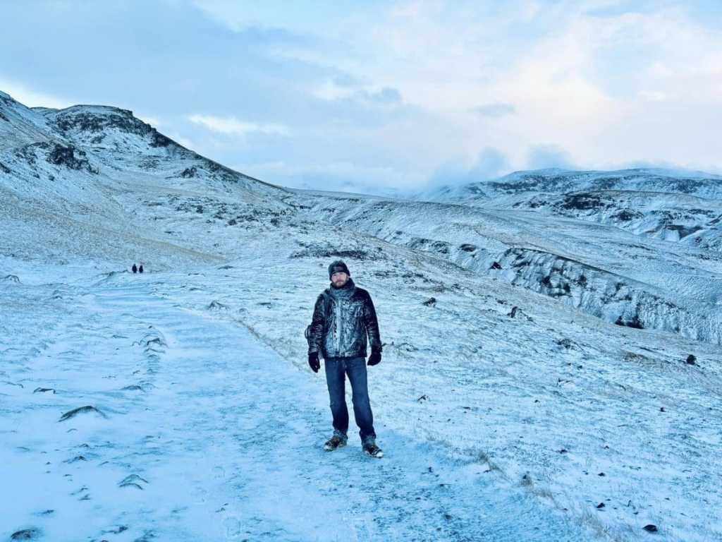A solo hiker wearing a patterned black jacket, beanie, and gloves stands on a winding, snow-covered trail that cuts through a vast geothermal valley in Iceland.