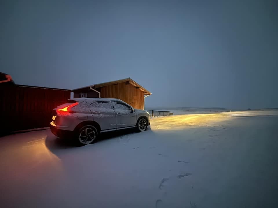 A white SUV parked in deep snow next to a wooden cabin at 9:00 AM in Iceland. The car's red taillights and bright yellow headlights are glowing against the dim, blue-toned morning light, illuminating the pristine, snow-covered ground. The surrounding landscape is a flat, white tundra stretching toward a soft, hazy horizon under a dark, moody sky.