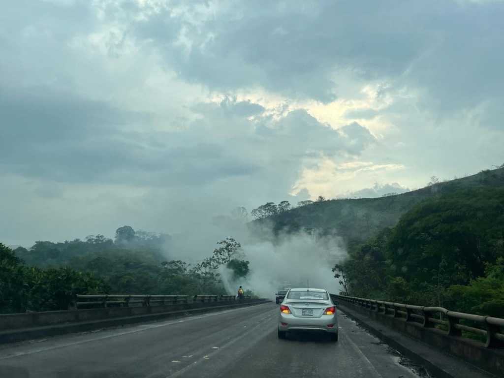 A silver car driving on a narrow bridge in Costa Rica, surrounded by thick white fog and lush green mountain forests under a heavy overcast sky.