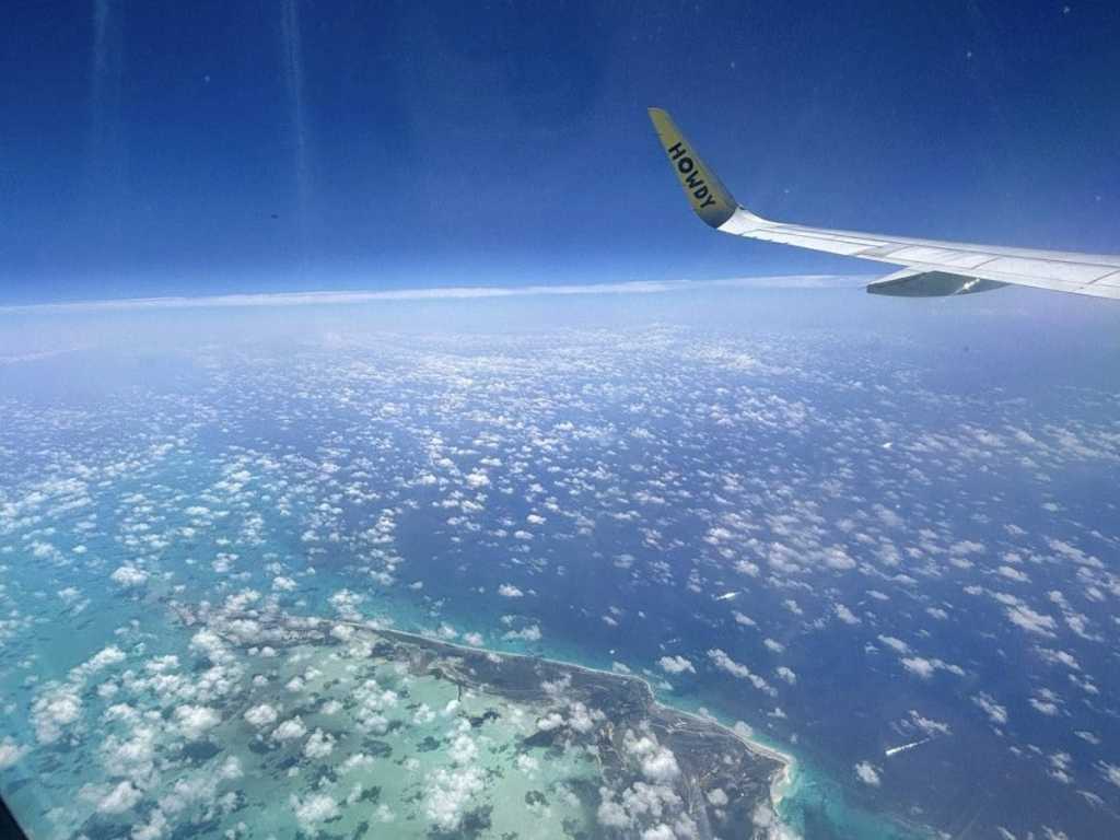A view from a Spirit Airlines plane window flying over turquoise ocean water and tropical islands, showing the yellow wing tip with "HOWDY" text, heading to Costa Rica.