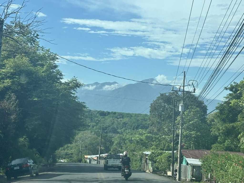 A view through a car windshield of a two-lane road in Costa Rica leading toward a massive, blue-toned mountain range under a bright sky. Lush green trees line the road, and a motorcyclist is seen riding ahead.