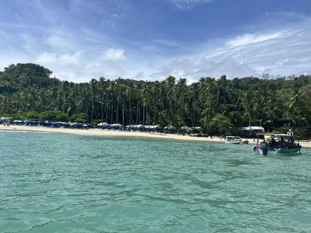 A view from the turquoise water looking toward the white sand shores of Tortuga Island, Costa Rica, lined with tall palm trees and blue beach umbrellas under a sunny sky.