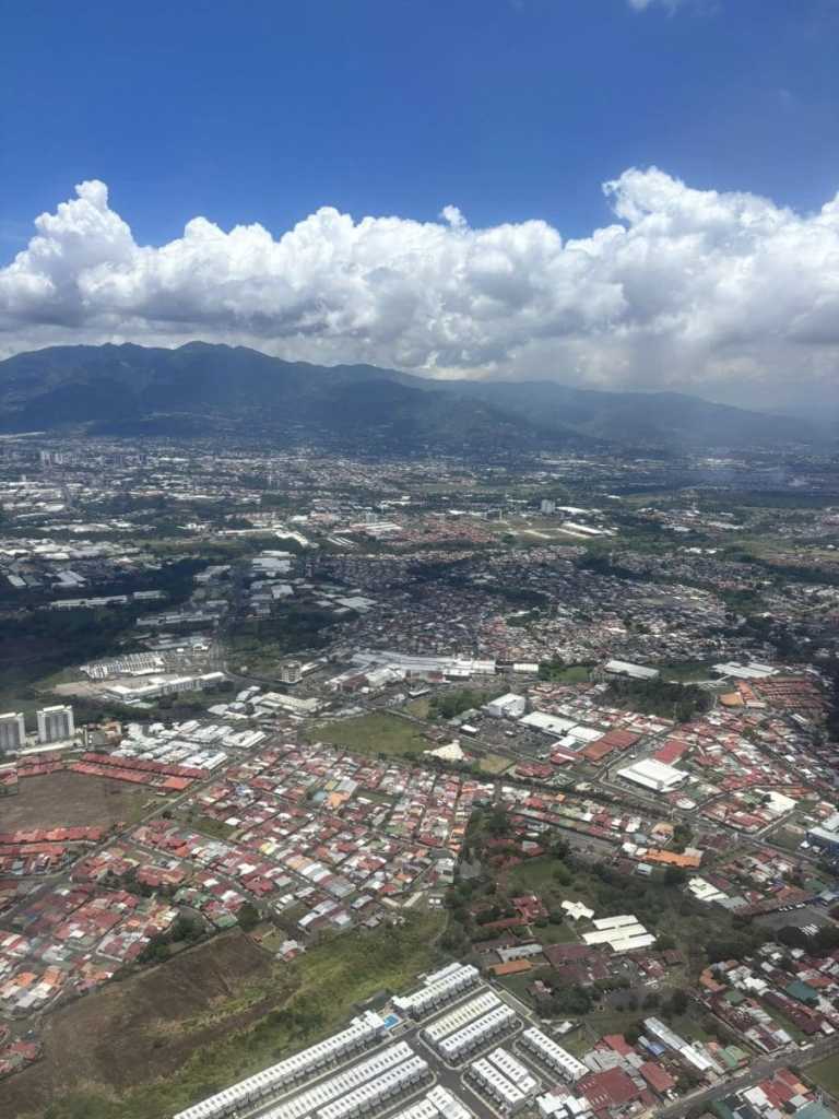An aerial view from a plane descending into San Jose, Costa Rica, showing the sprawling city landscape tucked between lush green mountains and white clouds.