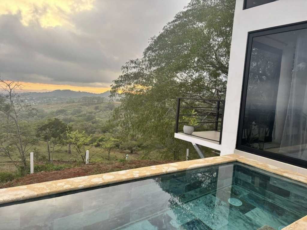 A luxury infinity pool at a modern Airbnb in Monte de Oro, Costa Rica, reflecting a glowing orange sunset over the lush green mountains and valley.
