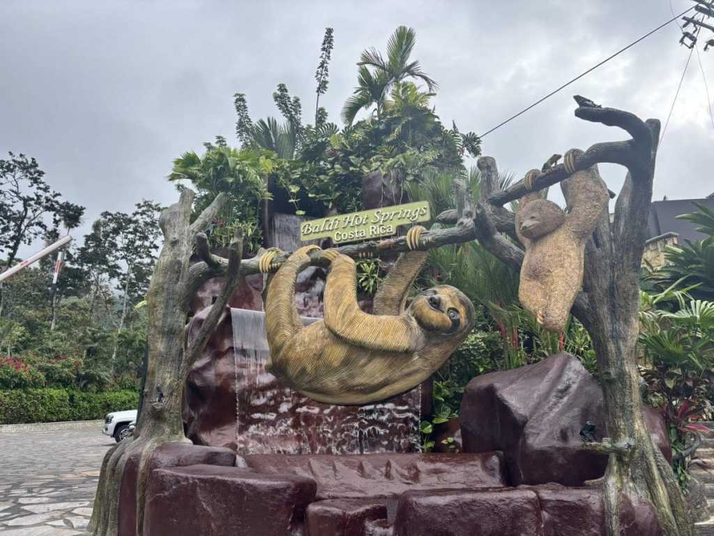 A decorative stone entrance for Baldi Hot Springs in Costa Rica, featuring a waterfall and large, realistic statues of sloths hanging from artificial tree branches against a backdrop of lush tropical plants.