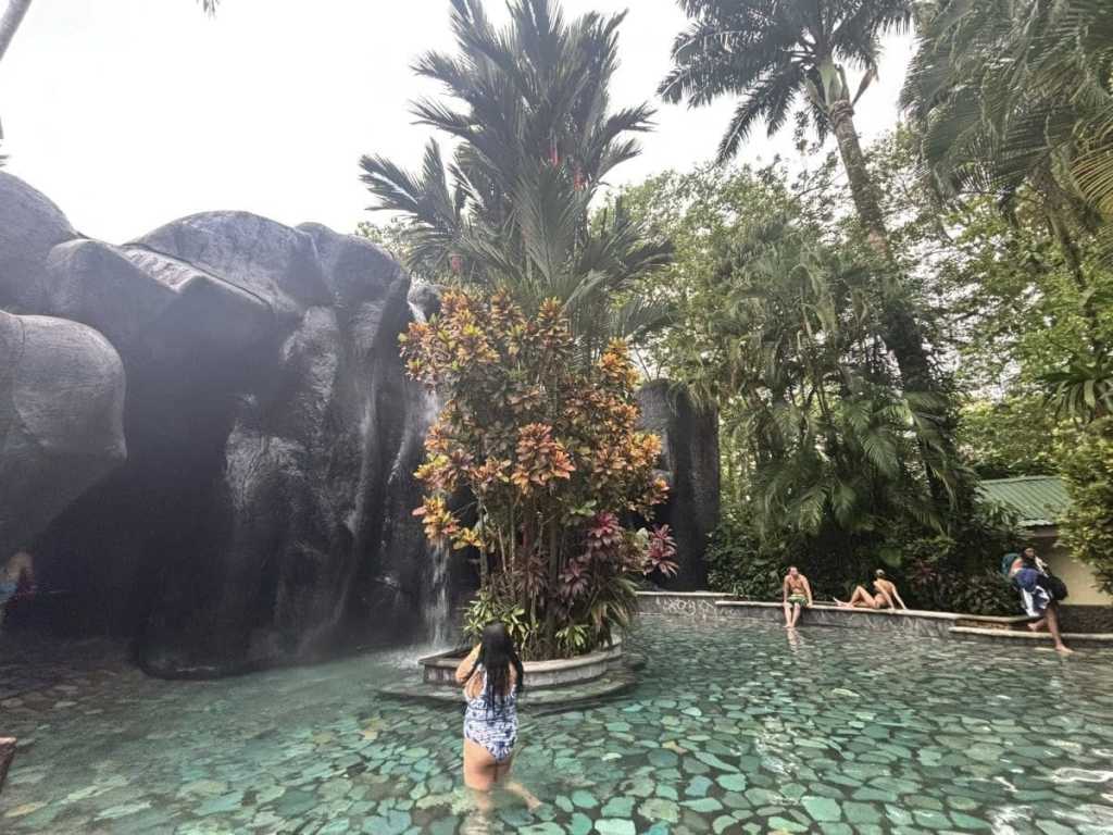 A woman in a blue patterned swimsuit wading through a crystal-clear thermal pool at Baldi Hot Springs in Costa Rica. The pool features a stone-tiled floor, a natural rock waterfall grotto, and is surrounded by vibrant tropical flowers and towering palm trees.