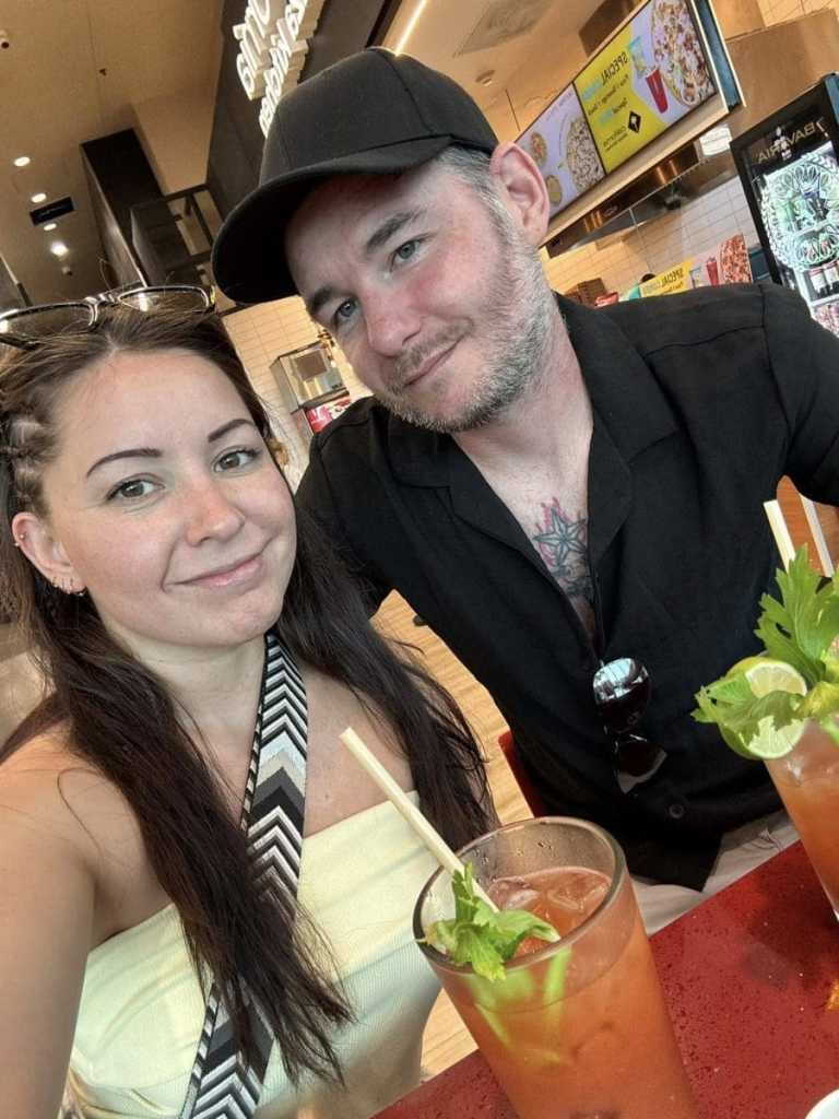 A selfie of a smiling woman and man sitting at a red airport restaurant table, enjoying Bloody Mary cocktails garnished with celery and lime before their flight.