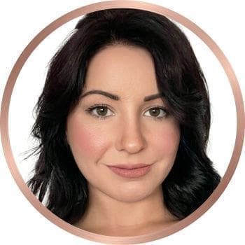 A circular professional headshot of Holly, a woman with dark wavy hair, centered within a thin rose gold metallic border against a clean grey background. Creator of the travel blog, "Holly Would Know."