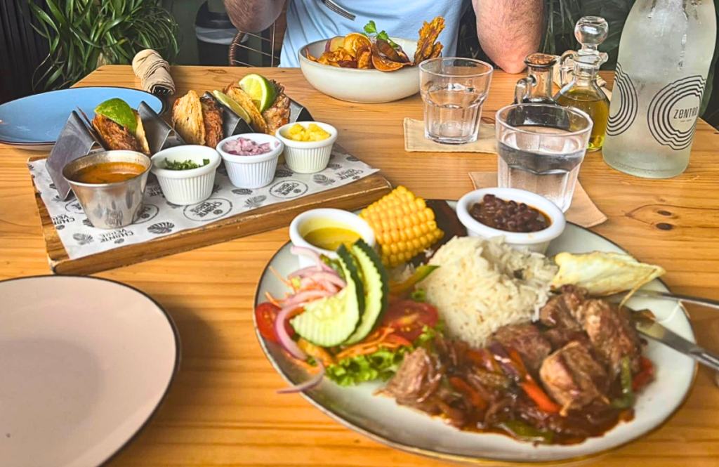 A top-down view of a colorful meal at El Chante Verde in Costa Rica. In the foreground, a plate holds steak sautéed with peppers, white rice, black beans, corn on the cob, and a fresh garden salad. In the background, a wooden board serves three tacos with small dipping bowls of salsa, onions, and cilantro. The setting is bright and tropical with warm wood tones and a glass water carafe.
